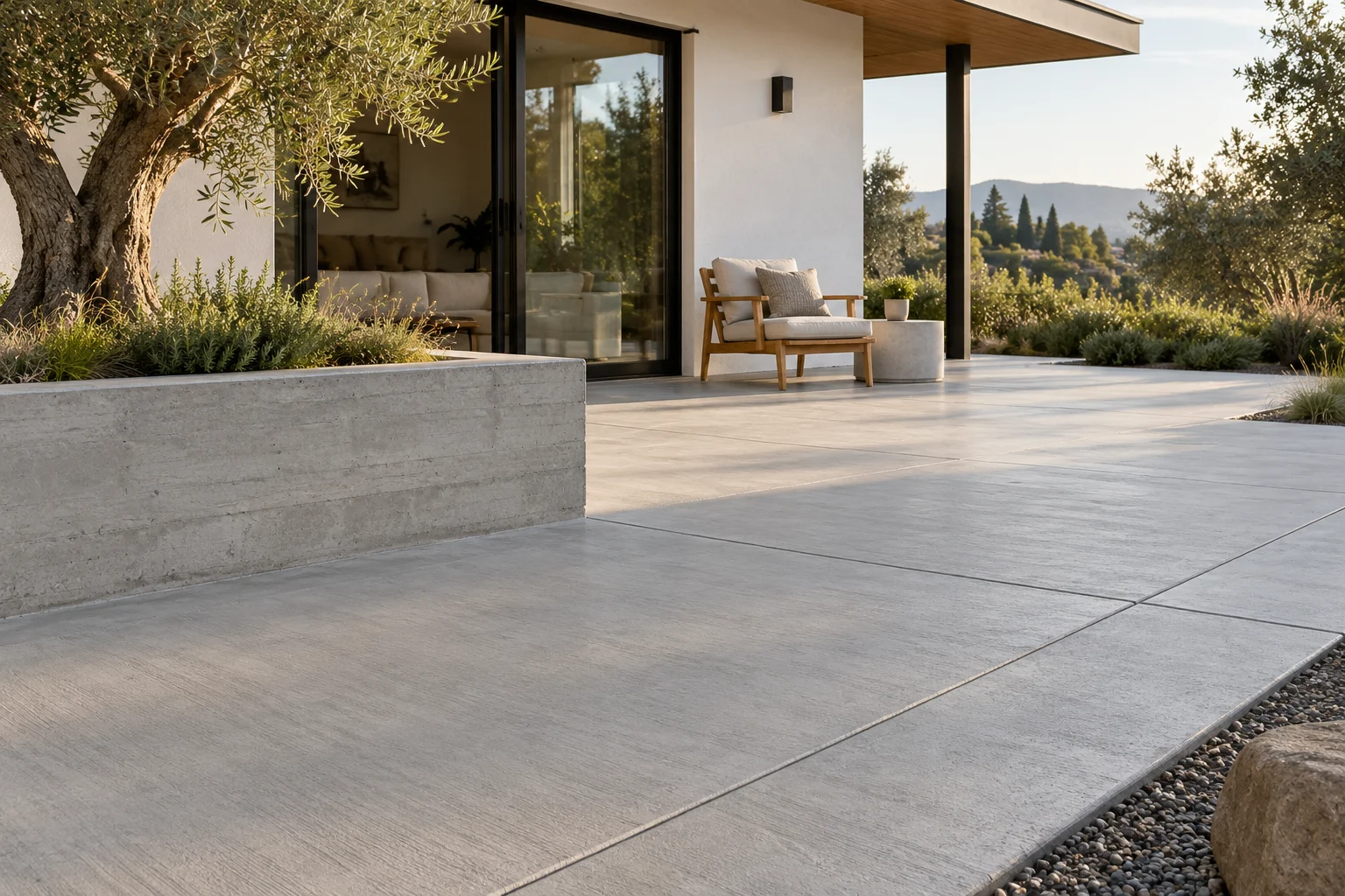 A concrete patio with joint lines extends past a raised planter and chair beside a modern house.